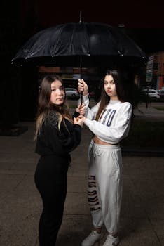 Two women in casual outfits holding a black umbrella outdoors during nighttime.