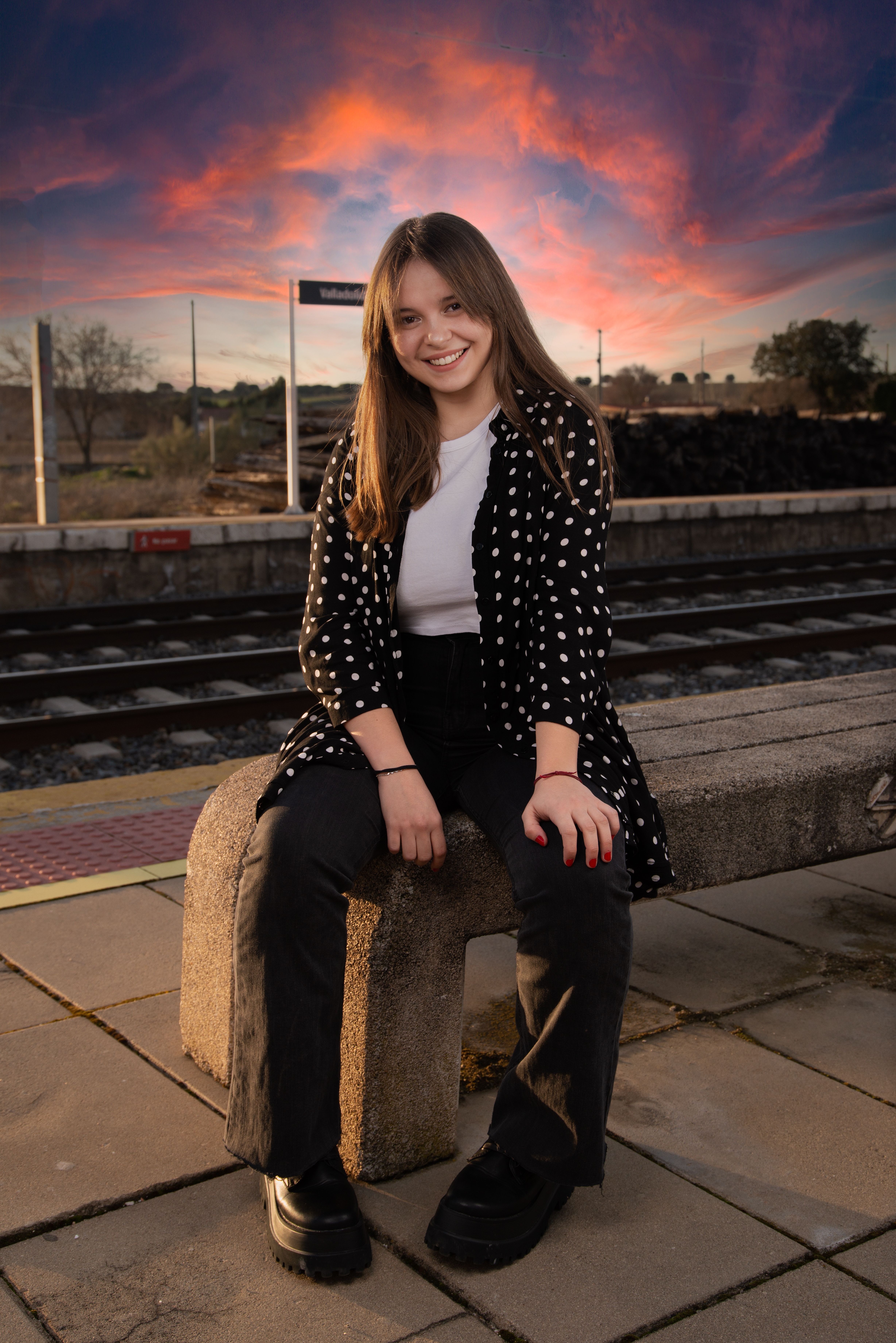 A cheerful young woman sits at a train station bench with a vibrant sunset sky.