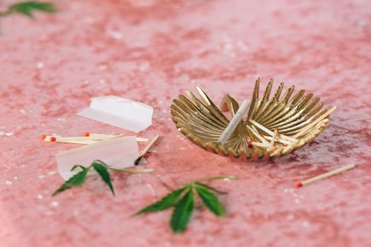 Gold ashtray holding cigarette and matches against a pink backdrop with green leaves.