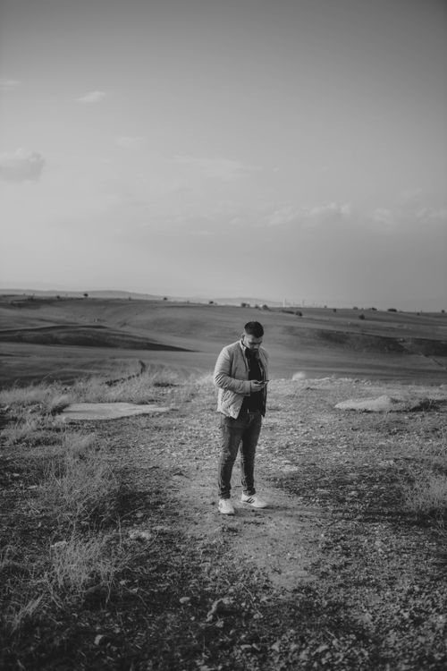 Free Full length of calm male wearing casual clothes walking in field while looking at screen of smartphone with hills on background Stock Photo