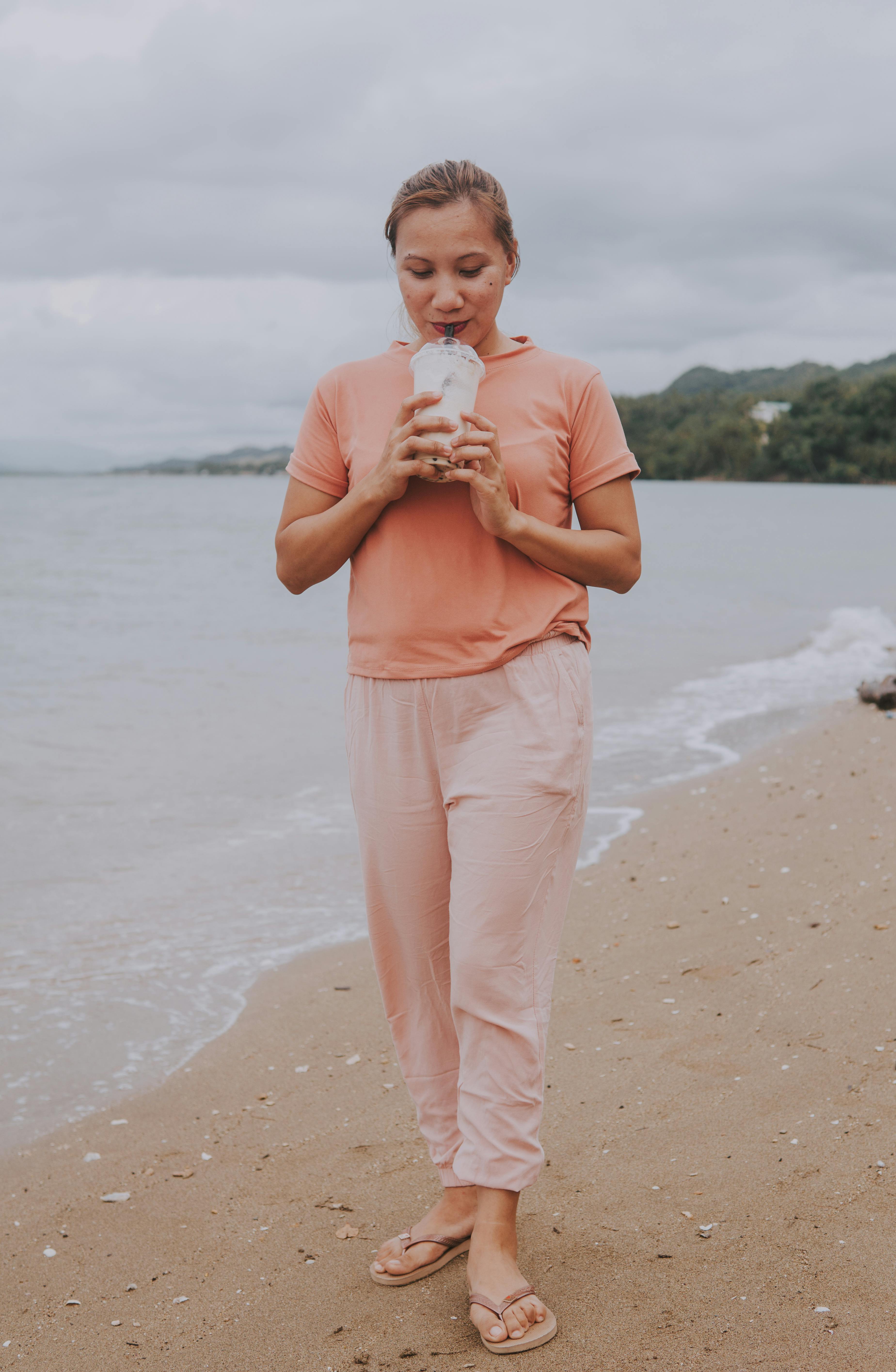 An adult woman enjoys a drink while standing on a sandy beach wearing a peach colored outfit.