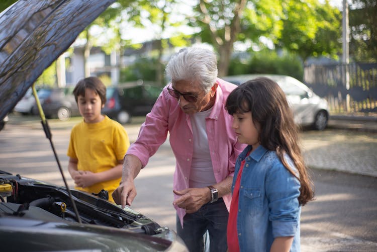 
An Man Looking At An Engine Of A Car With His Grandchildren