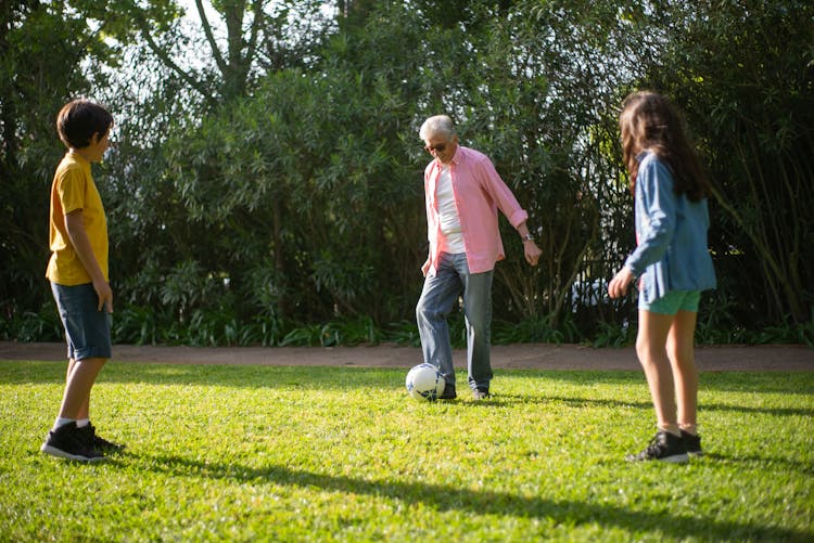 Elderly Man Playing Football With His Grandchildren On Green Grass