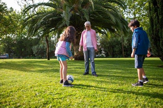 Grandfather and grandchildren playing soccer in a lush green park under a palm tree.