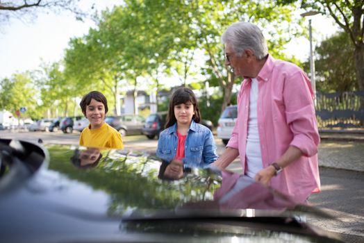 Grandfather and grandchildren interact by a car on a sunny day in Portugal.