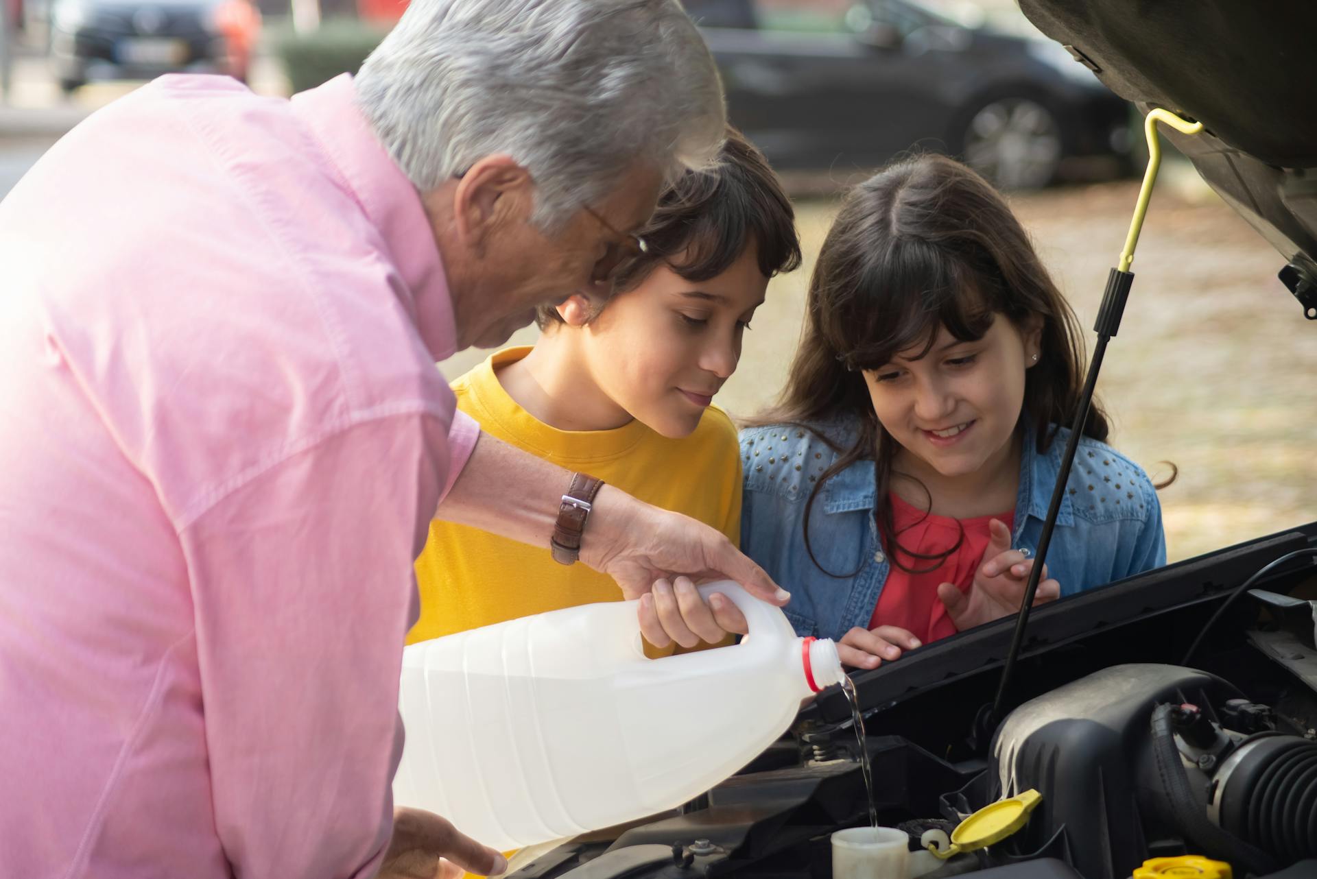 A grandfather guides his grandchildren in pouring fluid into a car engine outdoors.