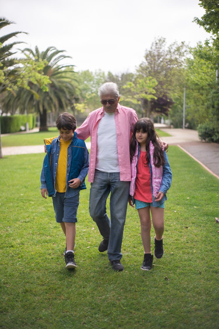 Elderly Man Walking On Green Grass With His Two Grandchildren