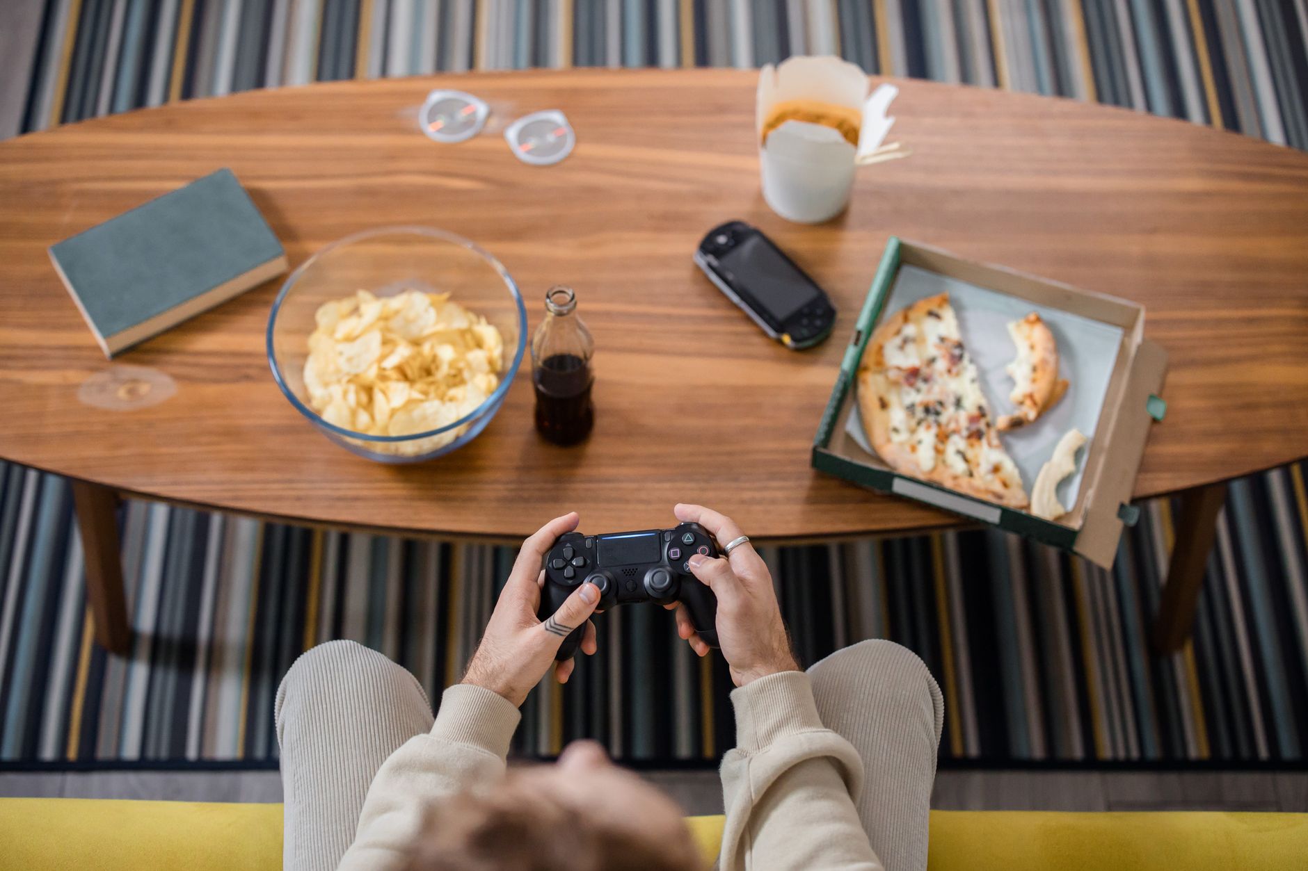 Overhead view of a person gaming with a controller, pizza, chips, and a drink on a table.