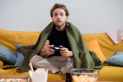 Man on couch gaming with controller, wrapped in blanket, offering cozy ambiance.