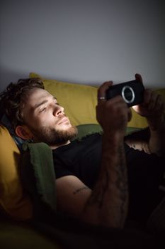 Bearded young man lying down playing handheld gaming console indoors, enjoying leisure time.