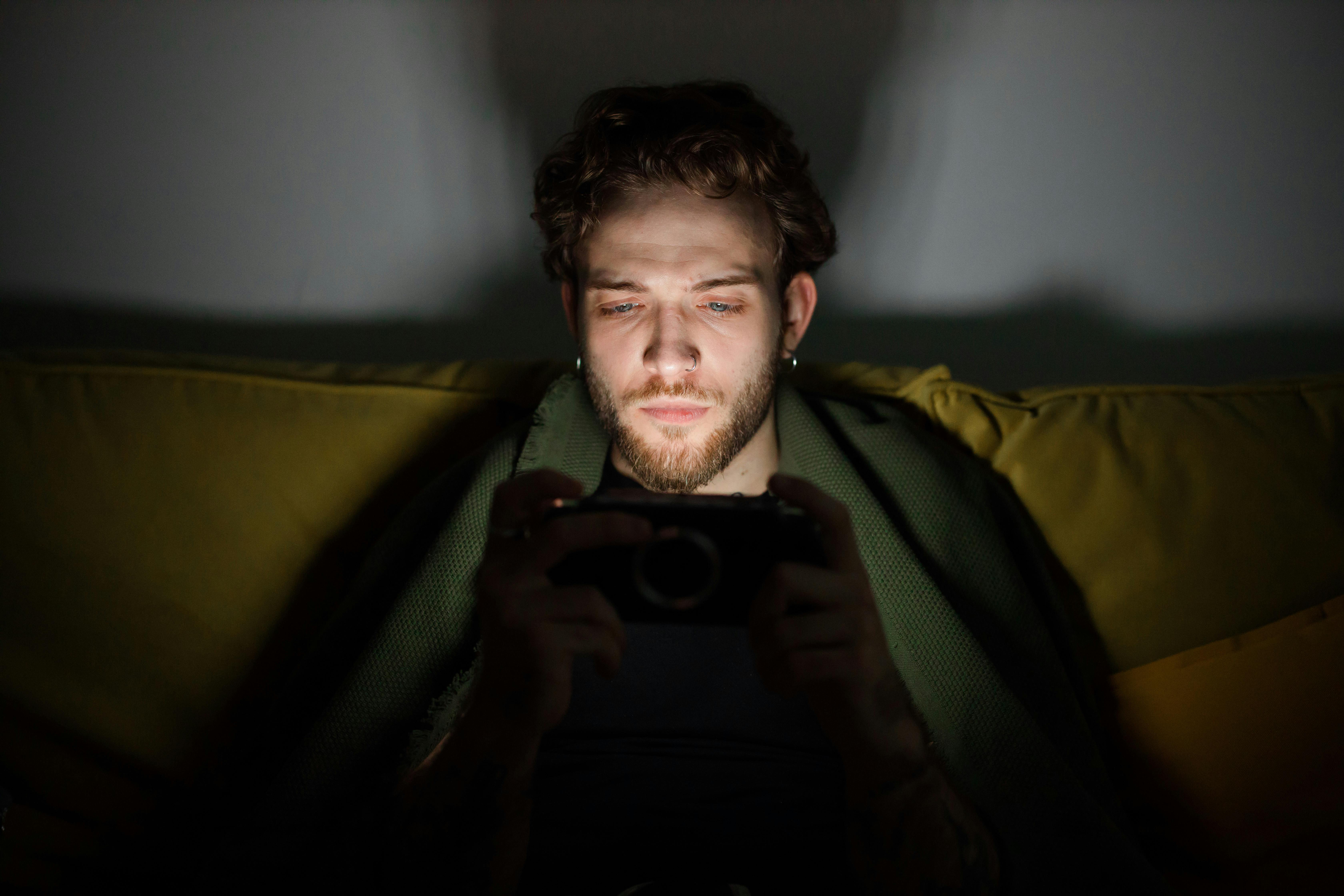 Free Focused young man looking at a smartphone screen, creating a moody atmosphere indoors. Stock Photo