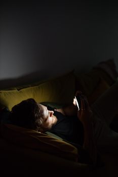 A man lounging on a couch playing games on his smartphone at night.