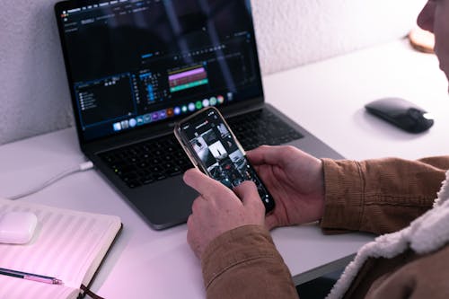 Man scrolling photos on smartphone while working on laptop