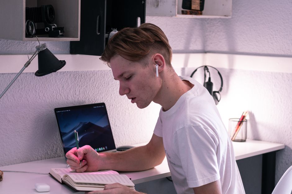 Side view of serious young man in TWS earbuds writing in notebook while studying on netbook at desk Side view of serious young man in TWS earbuds writing in notebook while studying on netbook at desk