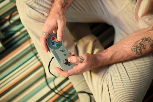 A close-up view shows tattooed hands using a retro game controller while sitting indoors.