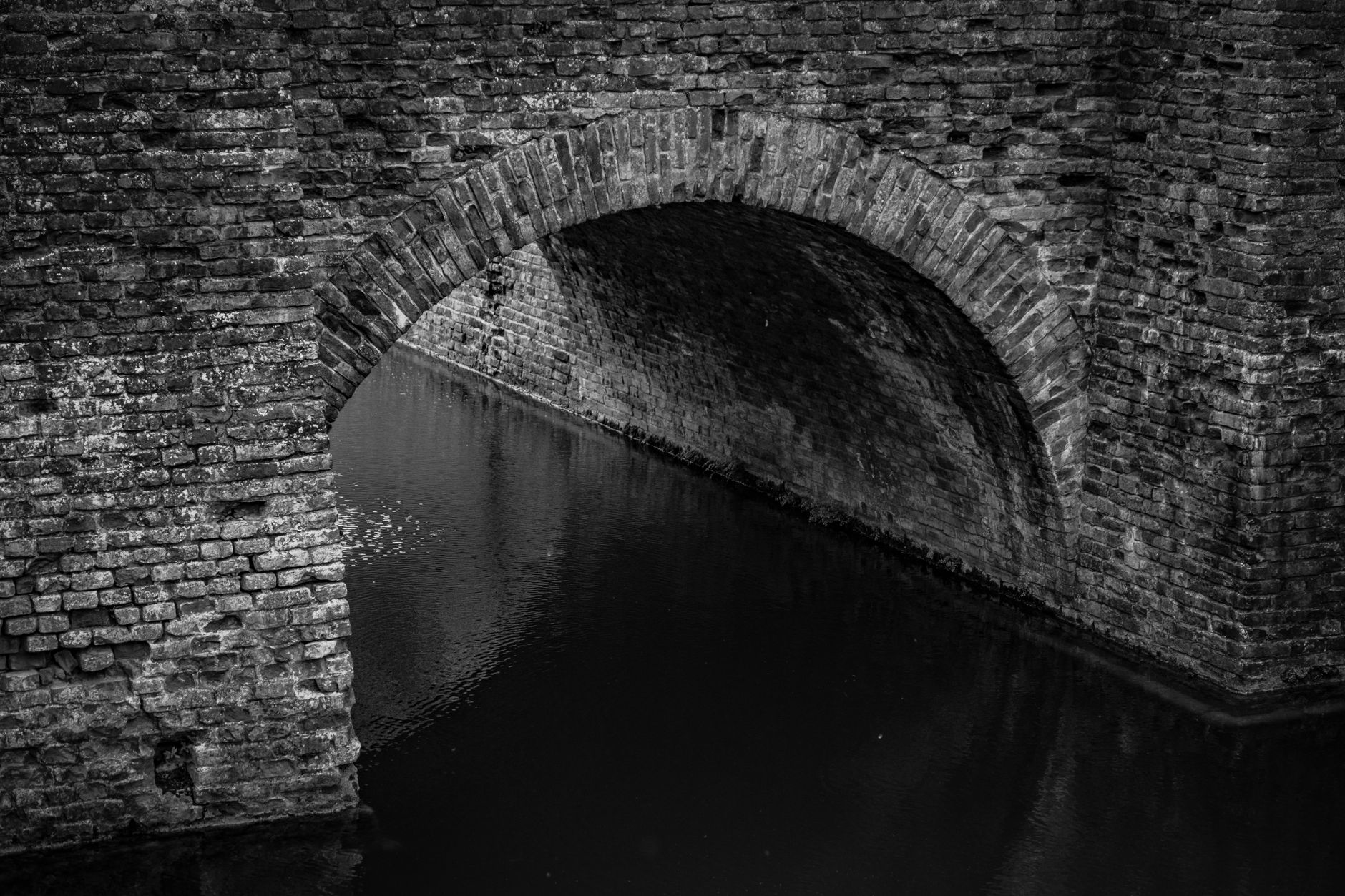 Black and white of arched aged brick wall bridge over rippling water of canal