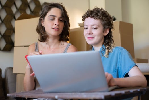 Two women working together on a laptop in a home office environment with boxes around.