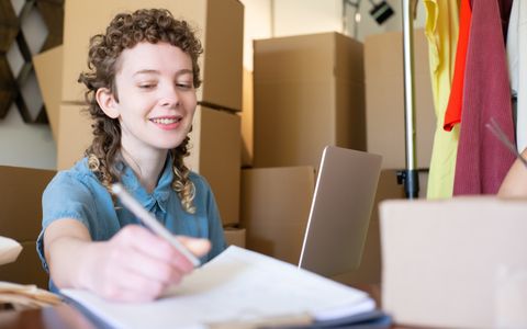 A small business owner writing notes at a desk surrounded by boxes
