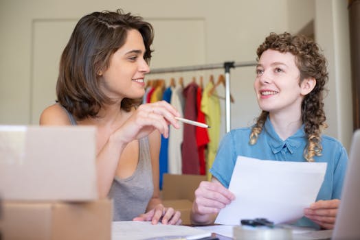 Two businesswomen discussing ideas in an office setting with clothing in the background.
