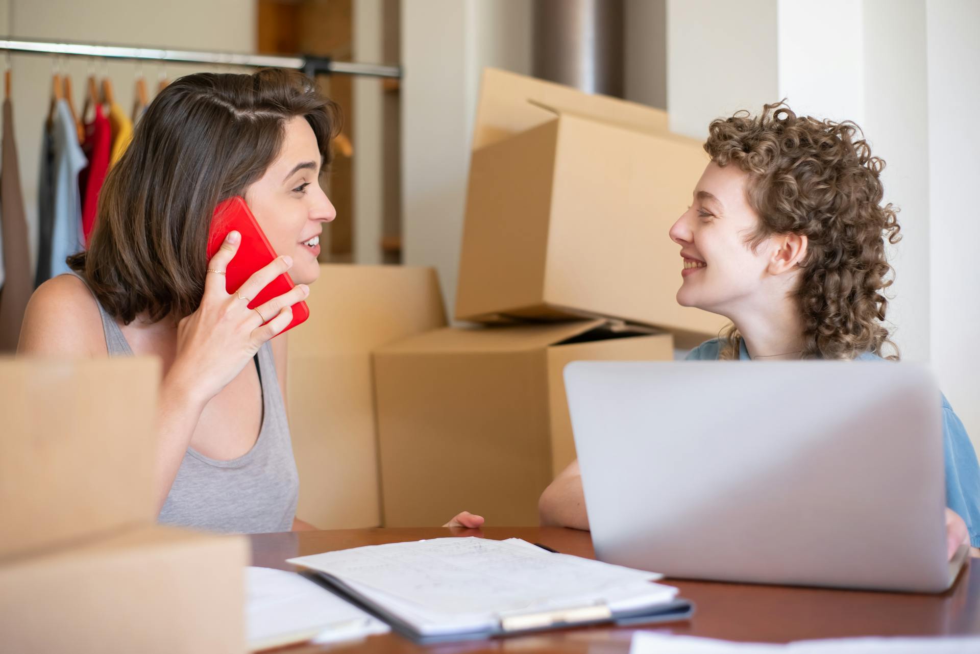 Two women managing their online clothing store using a laptop and phone, surrounded by boxes.