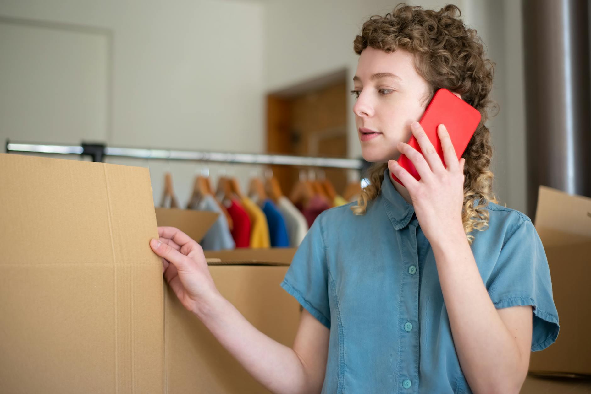 Young woman in blue shirt using phone while packing cardboard boxes indoors. Young woman in blue shirt using phone while packing cardboard boxes indoors.