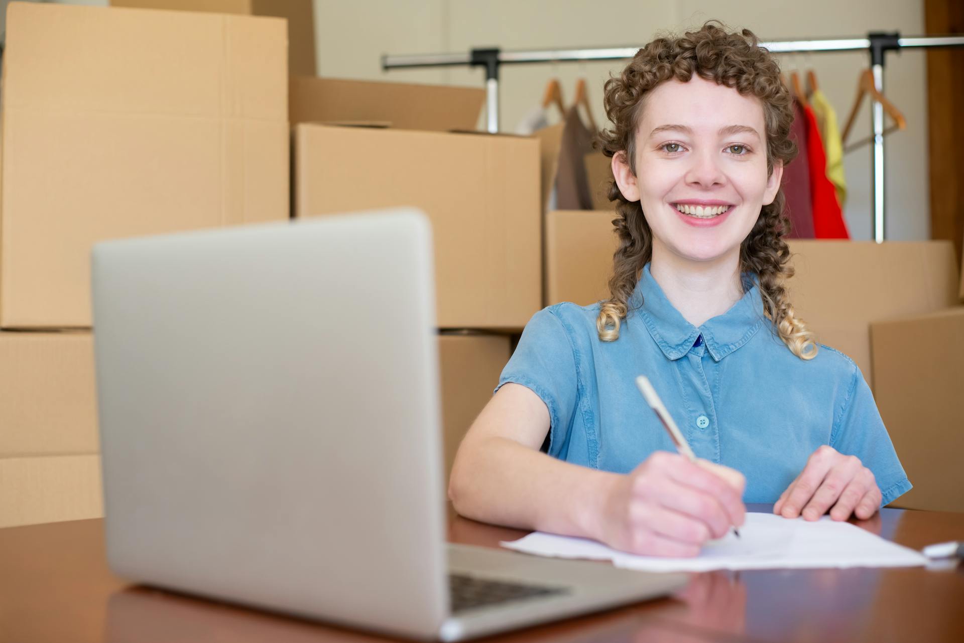 Cheerful woman writing notes at her home office, surrounded by boxes, symbolizing a startup or business setup.