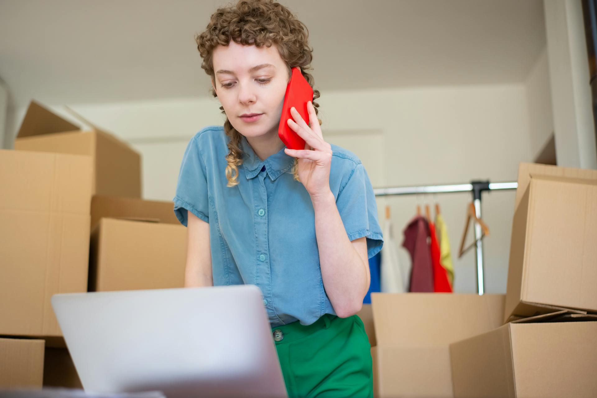 Focused woman multitasking with a phone and laptop in a home office surrounded by moving boxes.