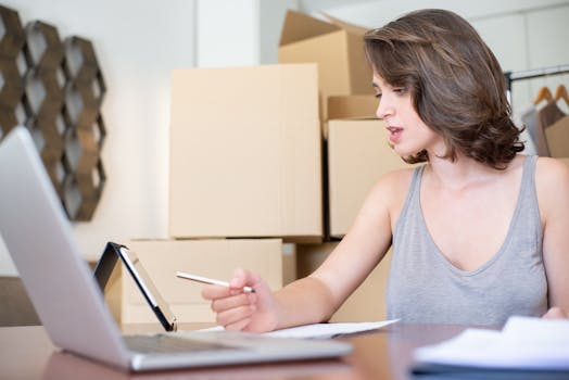 Woman running an online store, managing orders with a laptop and tablet, surrounded by cardboard boxes.