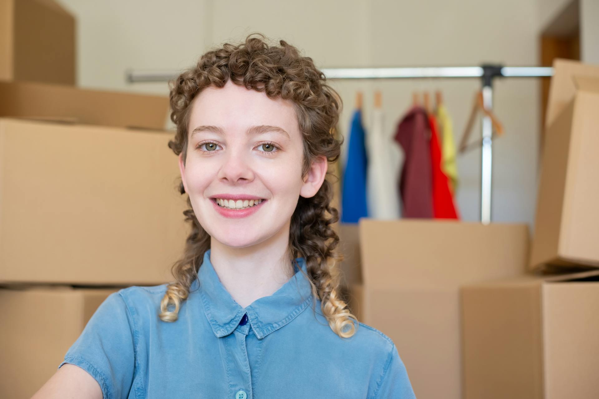 Happy young woman with curly hair standing among moving boxes indoors.