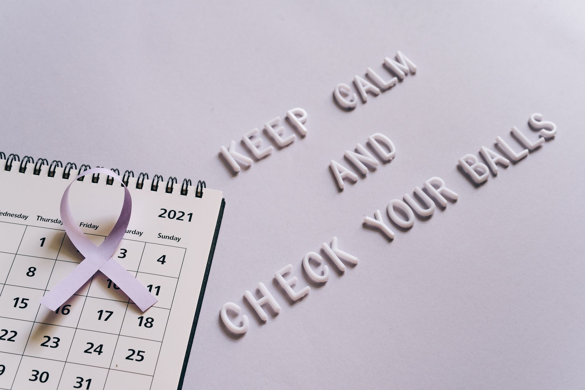 Flat lay image of a calendar with a purple ribbon and the text 'Keep Calm and Check Your Balls' for testicular cancer awareness.