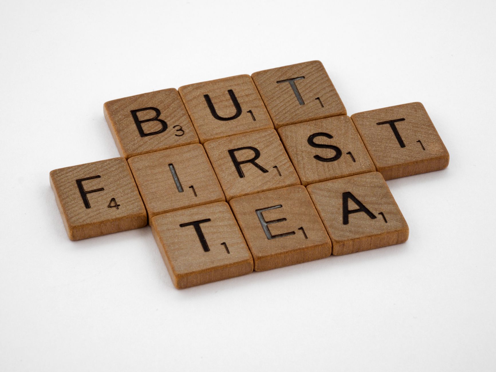 Creative arrangement of Scrabble tiles spelling 'But First Tea' on a white surface.