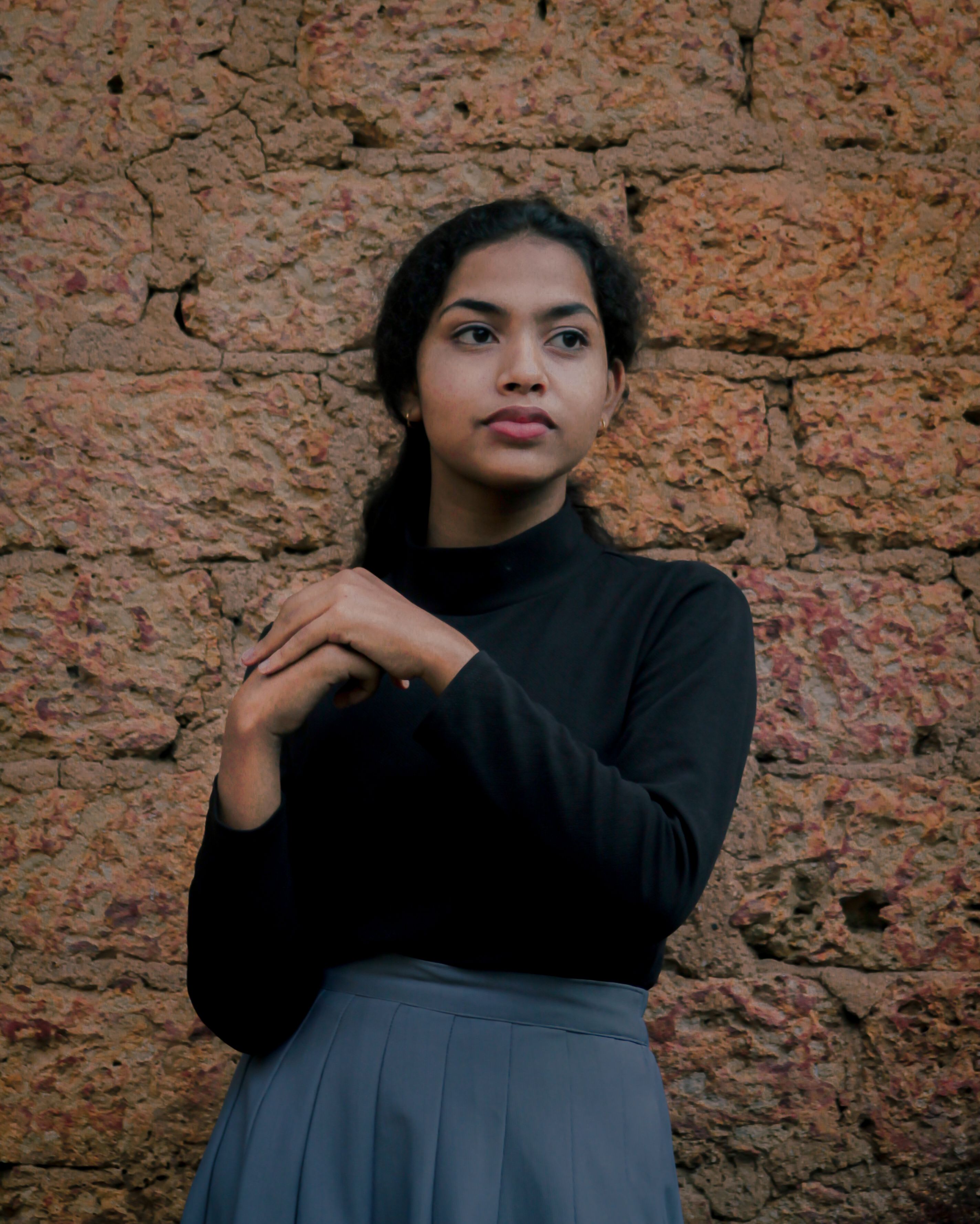 A thoughtful woman stands against a textured stone wall, gazing into the distance.