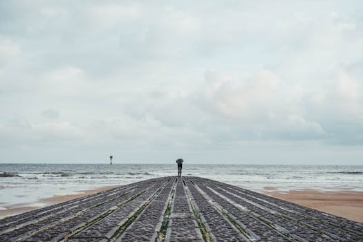 A lone person stands on a pier at Knokke-Heist beach, facing the sea under a cloudy sky.