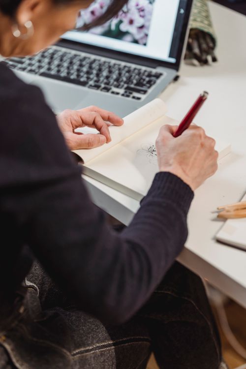 Free An artist is sketching in a notebook on a desk, accompanied by a laptop for inspiration. Stock Photo