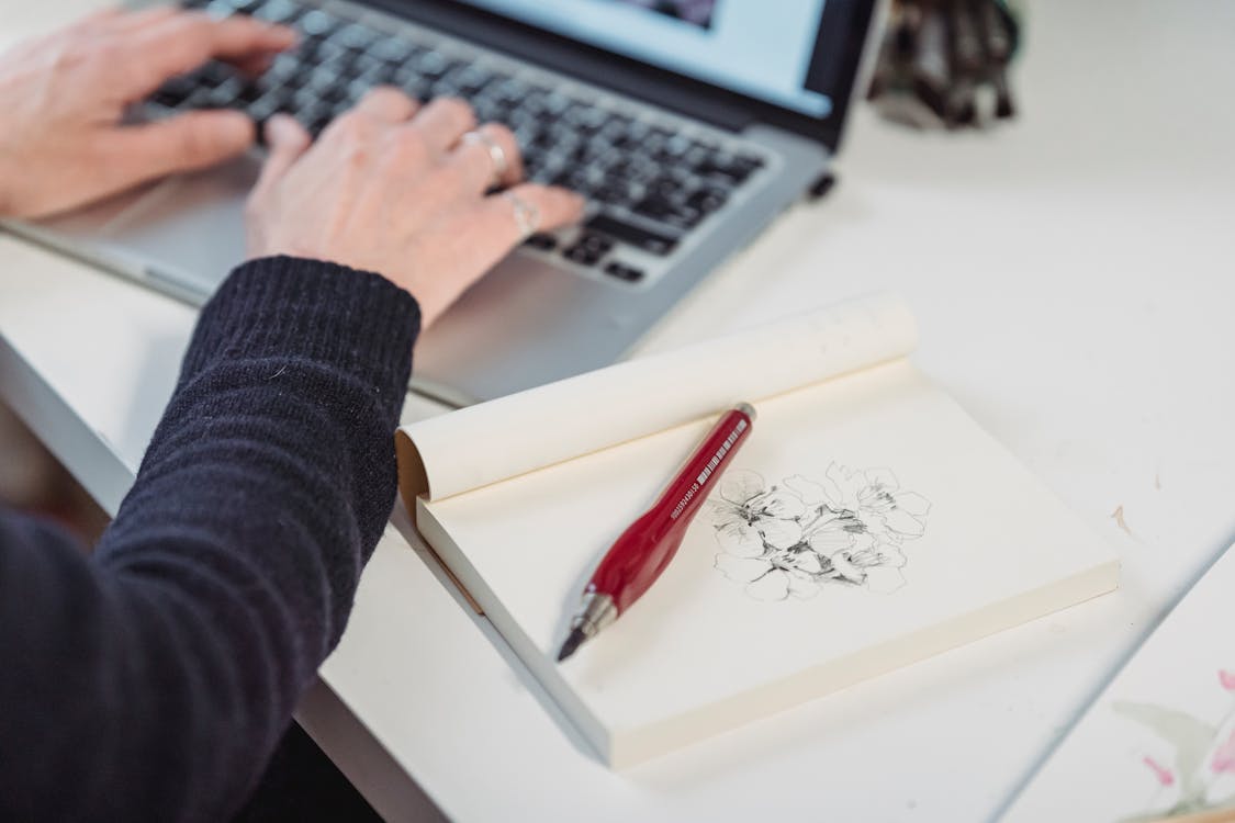 Free Close-Up Photo of Flowers Drawn in a Book beside a Person Using a Laptop Stock Photo