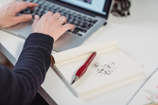 A person diligently typing on a laptop beside a sketchpad with flower drawings.
