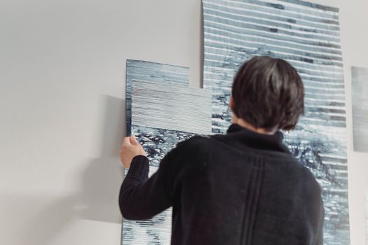 A woman in a dark sweater arranging abstract paintings on a wall, showcasing creative expression.