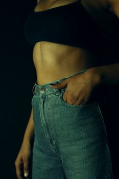 A woman in denim jeans and sports bra poses with hand in pocket.