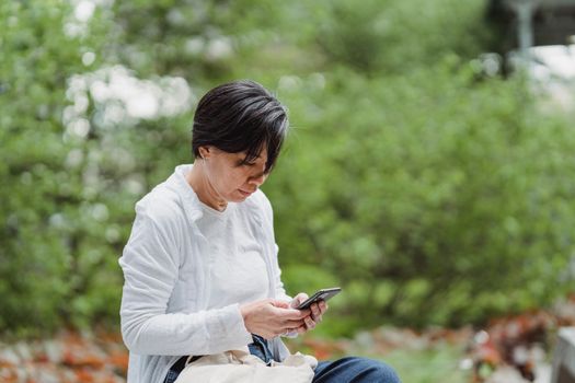 Woman sitting outdoors on phone, surrounded by nature. Captivated by screen.