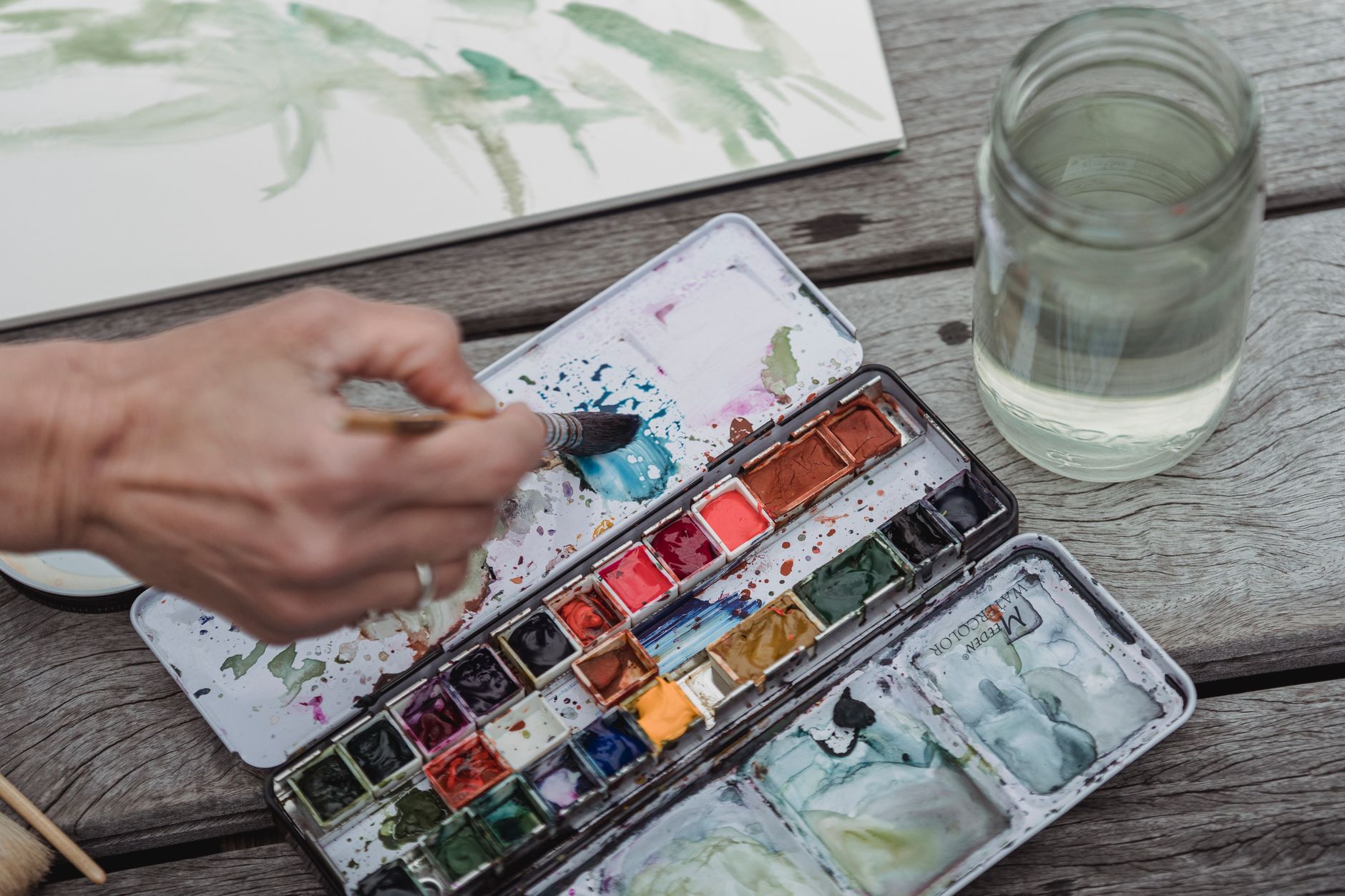 Hand using watercolour paints with a brush on a wooden surface
