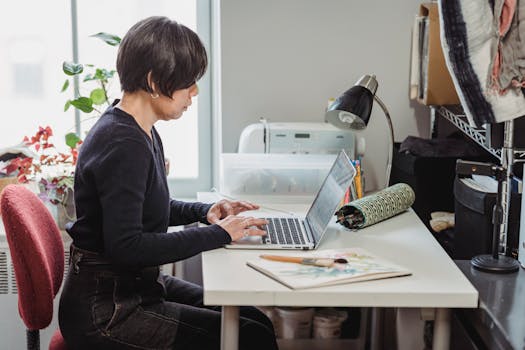 A woman working on a laptop in a cozy home studio environment.