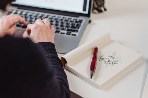 Free Hands typing on a laptop with a sketchbook and pen. Perfect for creative industry themes. Stock Photo