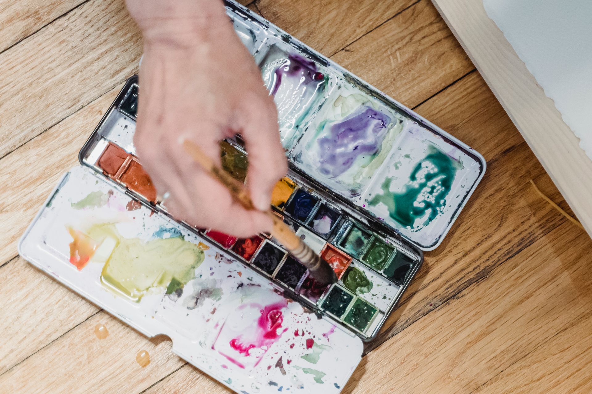 Close-up of a hand using a watercolor palette with vibrant pigments on a wooden floor.