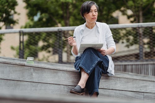 Woman in White Long Sleeve Shirt and Blue Denim Jeans Sitting on Brown Wooden Bench 