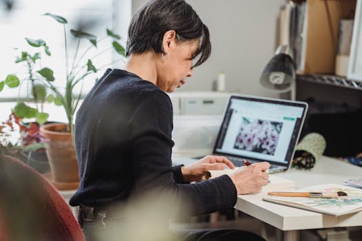 Woman focusing on creative work at a desk, surrounded by plants and a laptop, in a well-lit room.