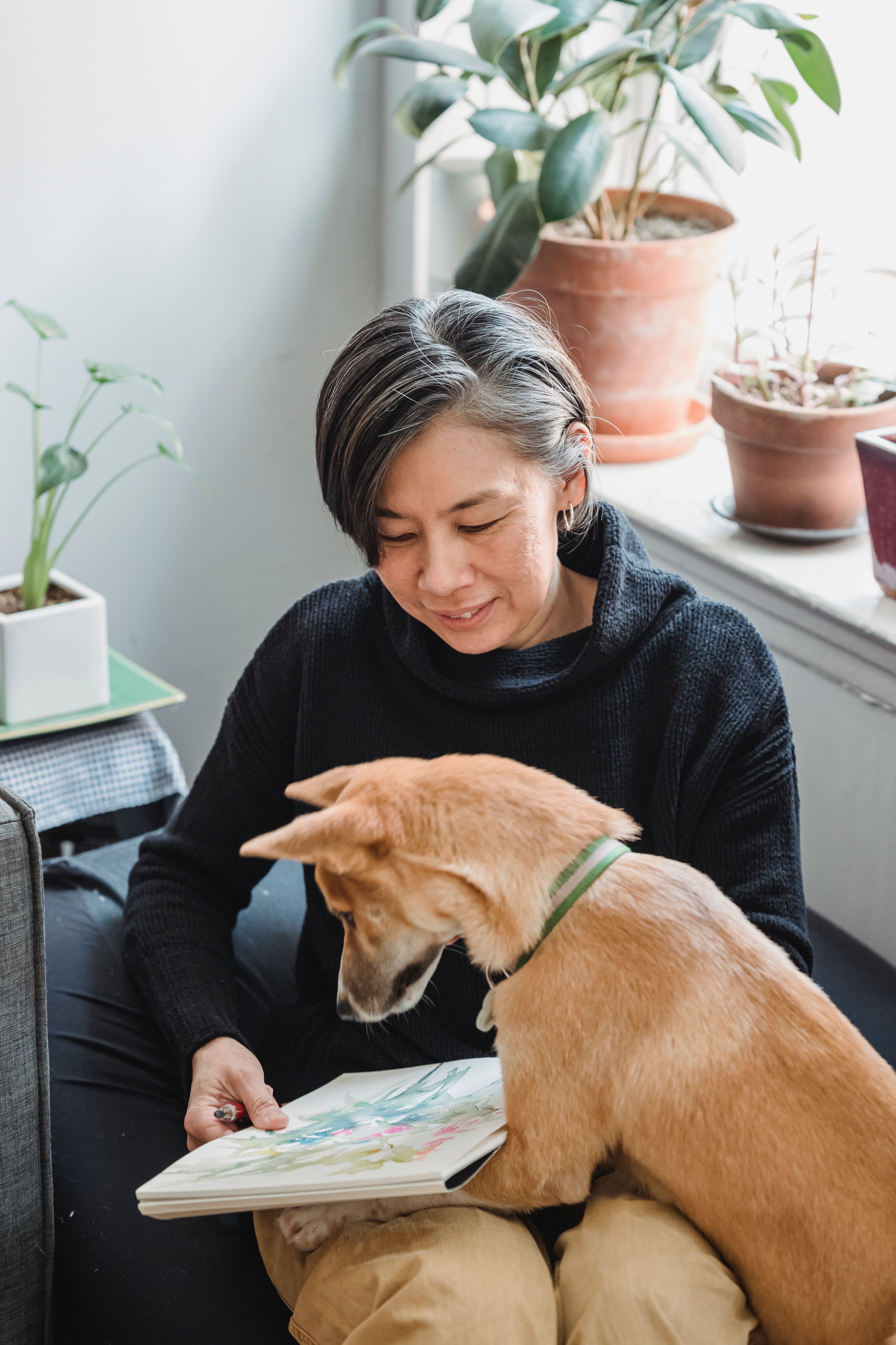 A woman enjoys sketching while her dog joins her on a cozy indoor sofa setting.