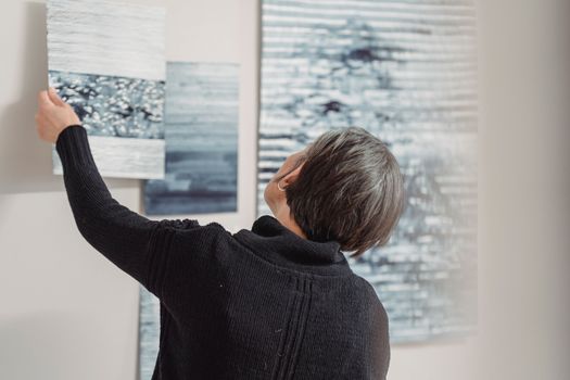 A woman arranges abstract art pieces on a gallery wall, showcasing modern creativity.