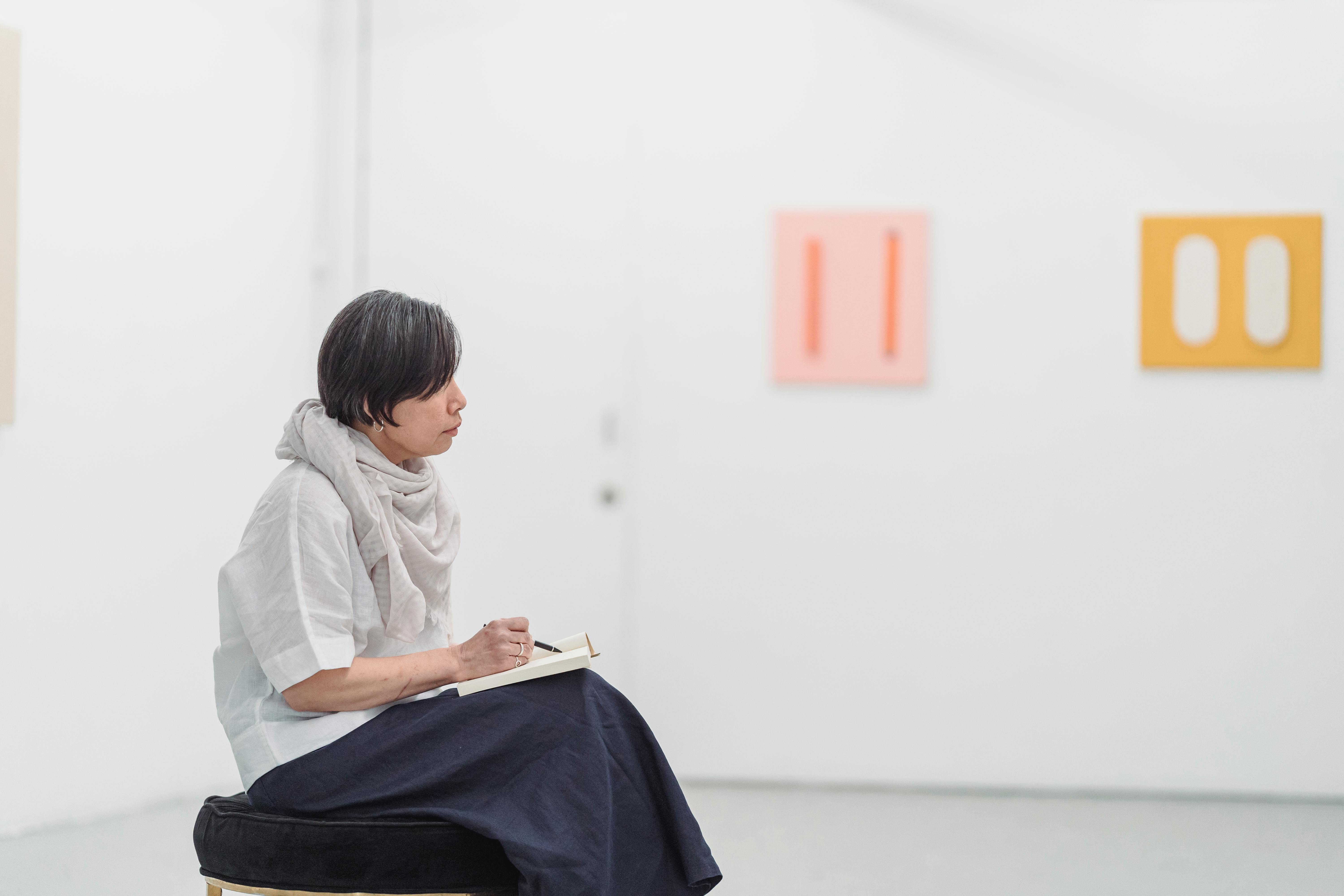 A woman thoughtfully sketches in a modern art gallery, surrounded by minimalist artwork.