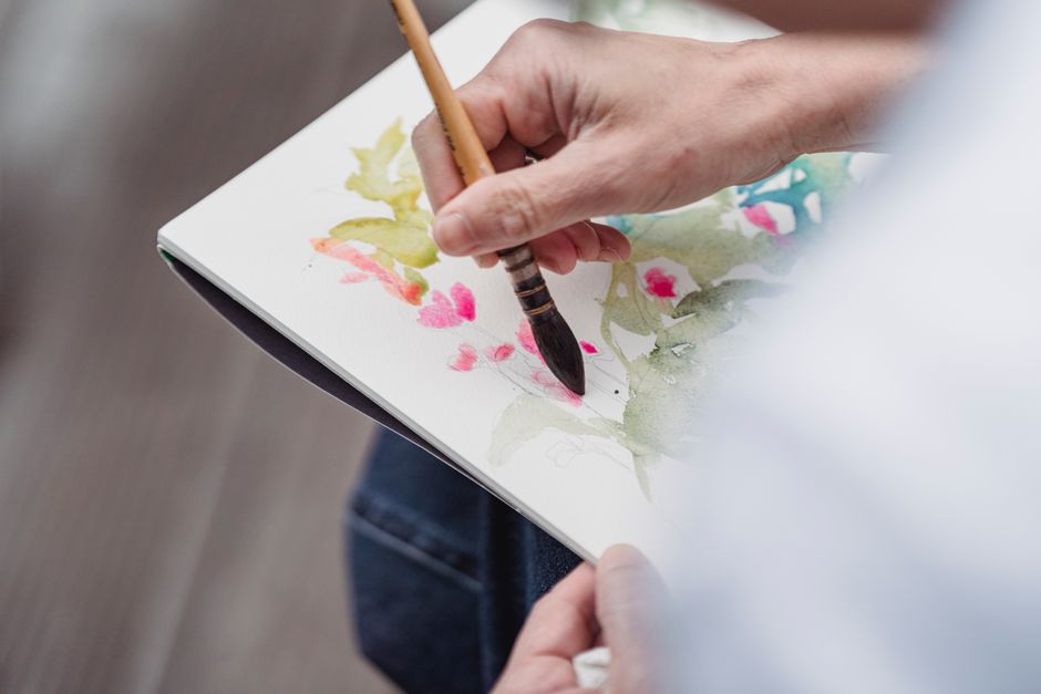 Artistic close-up of a hand using a brush to paint watercolor flowers.