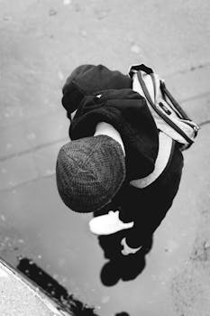 Black and white overhead shot of a person in a beanie and hoodie reflected in a puddle on a city street.